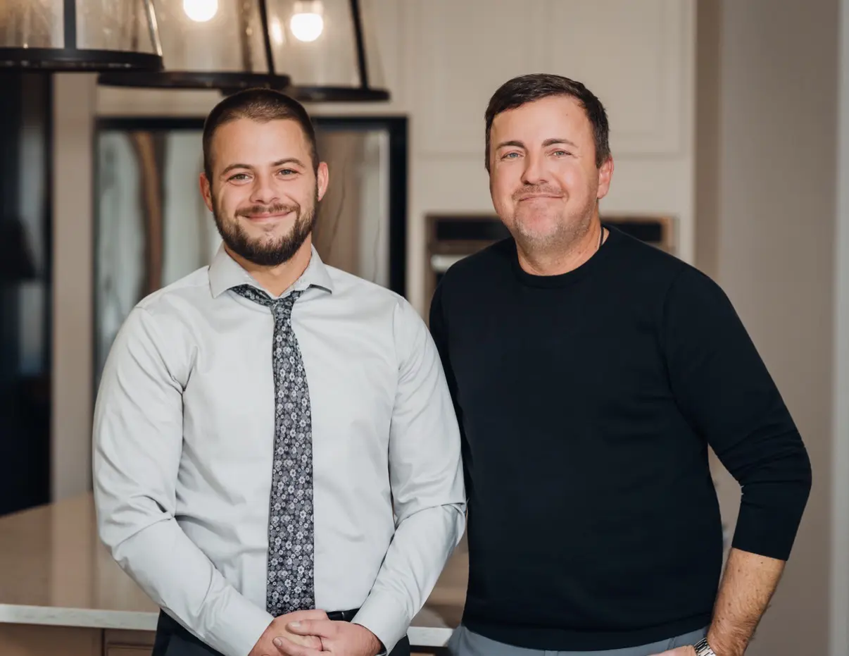 Two men smiling indoors, one in a shirt and tie, the other in a casual sweater.