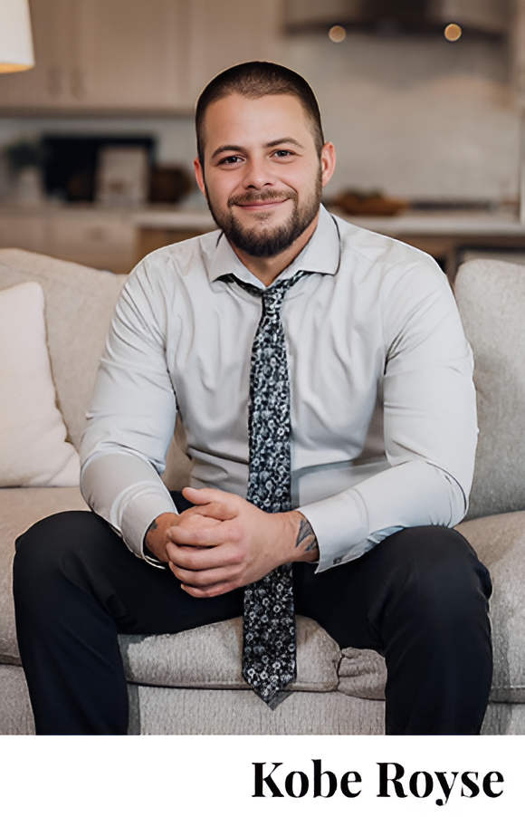 Smiling man sitting casually with hands clasped, wearing a white shirt and patterned tie.